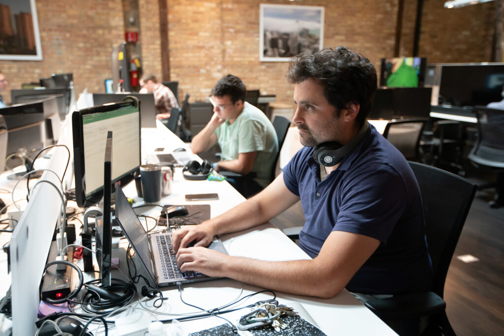 A male employee in a navy shirt and headphones on his neck works at a computer in an open office with other employees in the background.