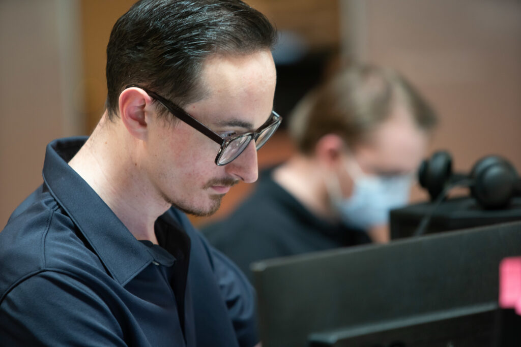A male Aspiritech employee with dark hair and glasses studies a computer screen.