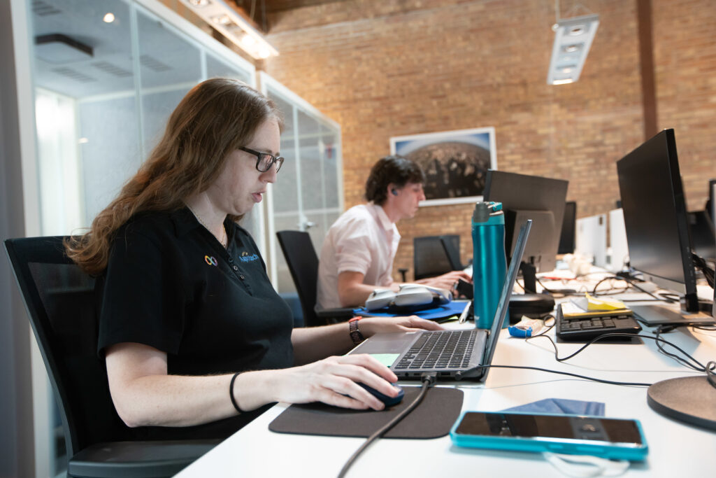 A female Aspiritech employee with long hair and glasses works at a laptop computer in an open office setting.