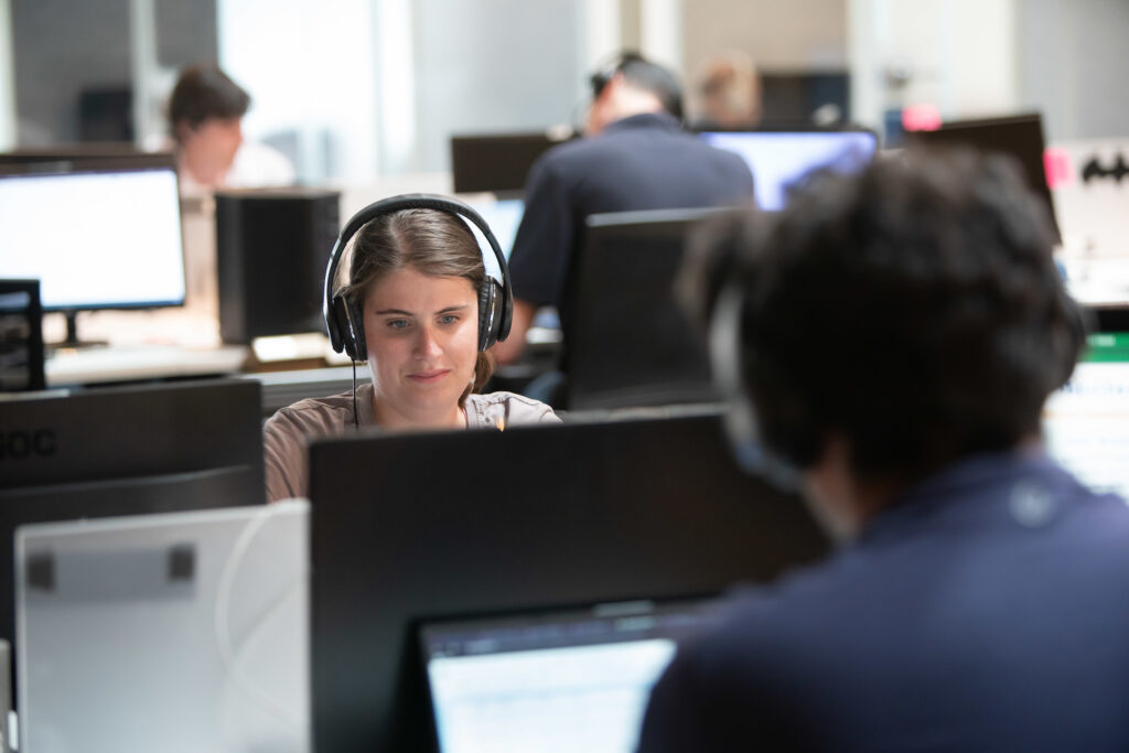 A woman with brown hair wearing headphones works at a desktop computer in an open office environment.