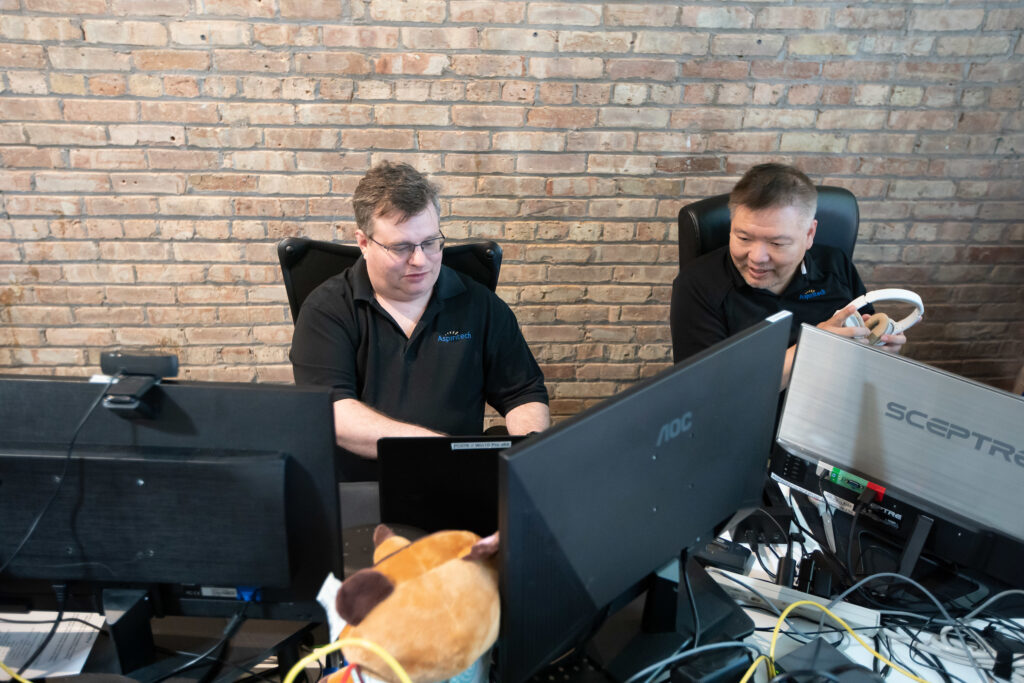 Two adult male employees work at computers in a brick office.