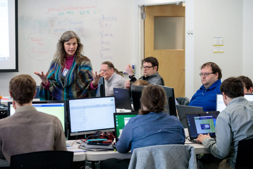 The mature white female trainer stands in front of six students demonstrating the QA testing lesson. Each student has a desktop in front of them.