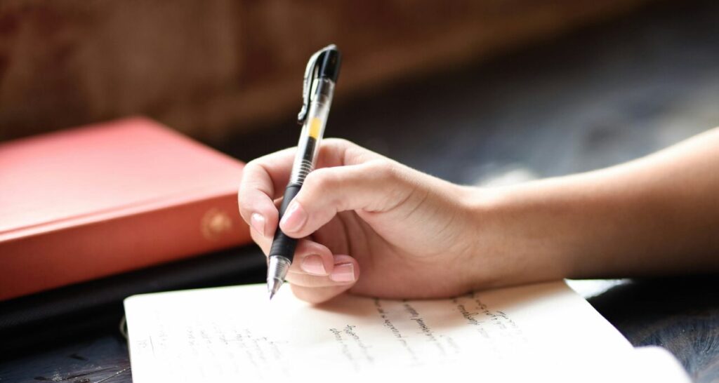 A brown hand holds a black pen over an open journal in front of a sunlight ledge topped with potted plants.