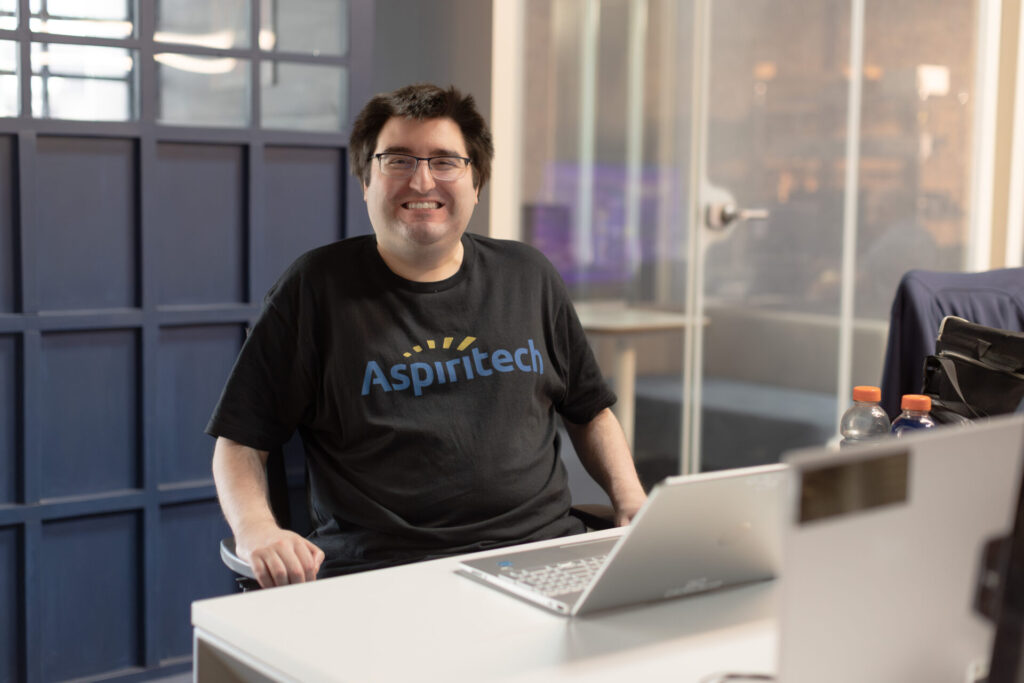 Alex Dudasik, a male Aspiritech employee, is wearing a black Aspiritech t-shirt and glasses smiles as he sits by his laptop on a long white desk.