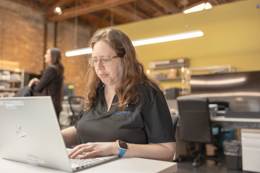 A female Aspiritech employee works at a laptop in an open office setting.